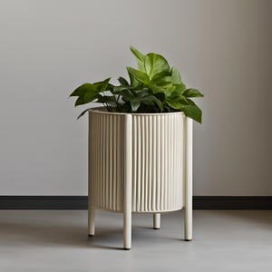 Op de afbeelding: Een crèmekleurige, cilindrische plantenbak met verticale geribbelde details. De plantenbak heeft vier poten en is gevuld met weelderige groene planten. Het ontwerp is modern en minimalistisch, geschikt voor binnen of buiten.