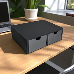 Puede incluir: Un organizador de escritorio negro con dos cajones grises. El organizador rectangular está sobre un escritorio de madera. Un portátil y una planta en maceta son visibles en el fondo.