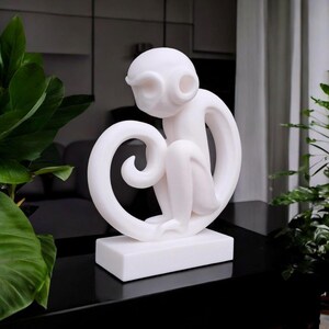 Op de afbeelding: Een witte apenbeeldhouwwerk met een gestileerd ontwerp. De aap zit op een rechthoekige basis, met zijn staart in een lus. Het beeldhouwwerk is gemaakt van een glad materiaal en is een decoratief item.