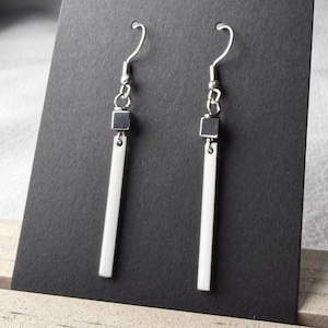 May include: A pair of minimalist earrings with a modern design. Each earring features a silver hook, a small square accent, and a long, white rectangular bar. The earrings are displayed against a dark background.