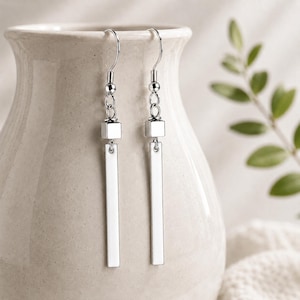 May include: A pair of silver-toned dangle earrings. Each earring features a small square, a chain, and a long, rectangular bar. The earrings are displayed against a neutral background with a blurred green plant.