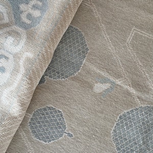 Pode incluir: Close-up de um têxtil bege e azul estampado. O tecido apresenta um padrão repetitivo de formas florais e geométricas estilizadas em tons de azul e bege. O têxtil está dobrado, revelando o verso com um design semelhante.