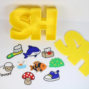 以下が含まれることがあります： 「SH」と書かれた黄色のブロック文字と、カラフルな漫画スタイルのステッカーのコレクション。ステッカーには、歯ブラシ、イルカ、ティッシュボックス、羊、日の出、魚、キノコ、茂み、青い靴が含まれています。