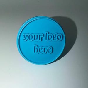 Può includere: Un oggetto rotondo blu brillante con le parole "your logo here" in rilievo. Il testo è in un carattere corsivo stilizzato. L'oggetto ha un bordo rialzato.