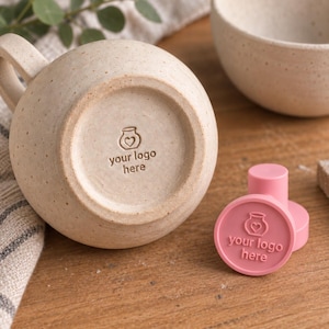 May include: A speckled beige ceramic mug with a handle, featuring a stamped logo of a jar with a heart on its base. Beside it sits a pink circular stamp tool that displays the same jar and heart logo with the text your logo here.
