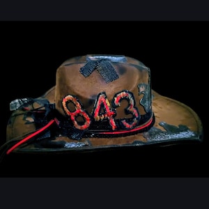 May include: Brown cowboy hat with the number "843" in red and black stitching. The hat has a distressed look with a black band and red accents. The background is black.