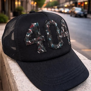 May include: Black baseball cap with a mesh back and a curved peak. The cap displays the number "404" in a colourful, patterned design on the front. The cap is resting on a stone surface, with a city street in the background.