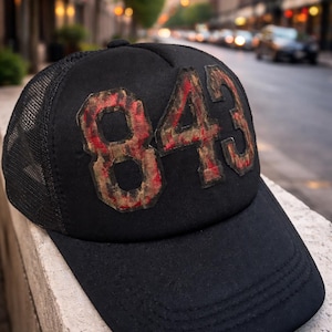May include: Black trucker cap featuring the number "843" in a distressed red and brown design. The cap has a mesh back and a curved peak. The hat is on a stone surface with a blurred city street in the background.