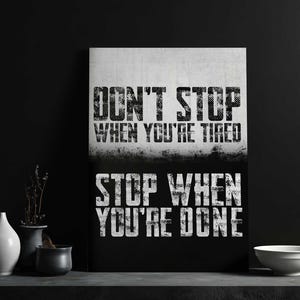 a motivational poster with the text "DON'T STOP WHEN YOU'RE TIRED" displayed prominently. The poster is set against a black background, and there are some decorative elements, such as vases and a bowl, placed in front of it.