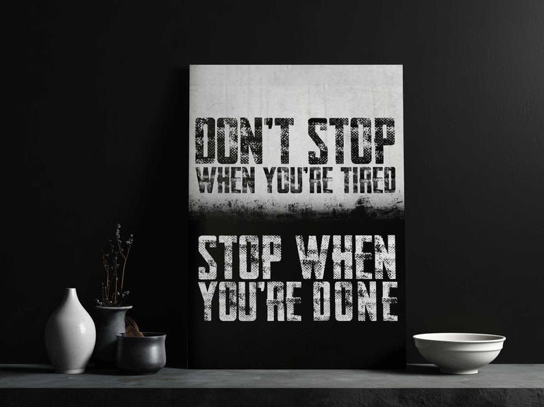 a motivational poster with the text "DON'T STOP WHEN YOU'RE TIRED" displayed prominently. The poster is set against a black background, and there are some decorative elements, such as vases and a bowl, placed in front of it.