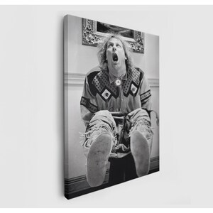 May include: A black and white canvas print featuring a person with long hair, mouth open in an upward expression, wearing a patterned long-sleeve shirt with a bolo tie and fringed moccasins. The person is positioned in front of a wall with a framed picture.