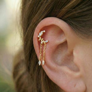 Op de afbeelding: Goudkleurige oorbel met een halve maan en een hangend kettingontwerp. Het ooraccessoire is versierd met heldere, sprankelende edelstenen. De sieraden worden op het bovenste oorkraakbeen gedragen.