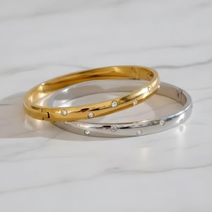 May include: Two elegant bangles. One is gold-toned, the other is silver-toned. Both are adorned with evenly spaced, sparkling clear crystals. The bangles are displayed on a white marble surface, reflecting light.