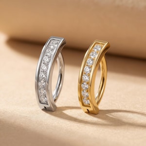 May include: Two curved barbell body jewelry pieces, one in silver-tone metal and one in gold-tone metal. Each piece features a channel setting containing seven round, clear, faceted stones arranged in a vertical row along the curved bar.