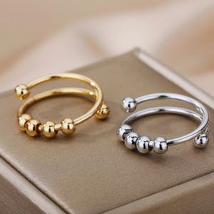 Pode incluir: Dois anéis ajustáveis, um dourado e outro prateado, cada um com um design de banda dupla. O anel dourado tem contas douradas, e o anel prateado tem contas prateadas. Os anéis são exibidos em uma superfície marrom.