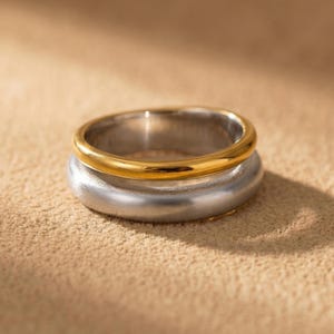 Może przedstawiać: Dwukolorowy pierścionek ze złotą obrączką na srebrnej obrączce. Pierścionek jest umieszczony na neutralnym tle, z miękkim oświetleniem i cieniami. Pierścionek to biżuteria.