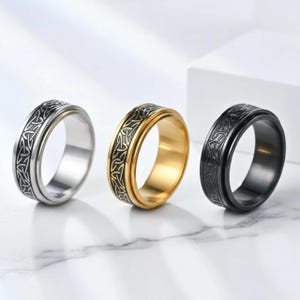 Peut inclure: Trois bagues en acier inoxydable avec un motif de nœud celtique. Une bague est argentée, une est dorée et une est noire. Les bagues ont une bande centrale rotative avec le motif de nœud celtique.