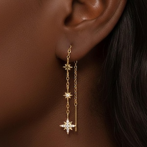 Peut inclure: Boucles d'oreilles en forme d'étoiles dorées avec un design de chaîne délicate. Les boucles d'oreilles comportent trois breloques en forme d'étoile, la dernière étant ornée de petites pierres scintillantes.