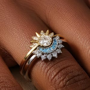 Op de afbeelding: Close-up van drie gouden ringen. De bovenste ring heeft een zonnesterontwerp met heldere en blauwe edelstenen. De middelste ring heeft een gouden band en de onderste ring heeft een zilveren band. De ringen zitten om een vinger.
