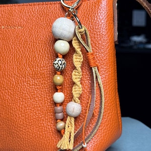 May include: Orange leather handbag with a decorative beaded keychain. The keychain features wooden beads in various sizes and colors, macrame detailing, and leather accents. The bag has a gold-colored clasp.