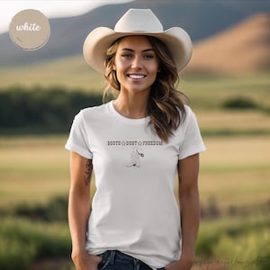 Könnte beinhalten: Weißes T-Shirt mit dem Text "BOOTS DUST FREEDOM" und einer Cowboy-Stiefel-Grafik. Das Shirt hat einen Rundhalsausschnitt. Im Hintergrund ist eine Person mit Cowboyhut und eine natürliche Landschaft zu sehen.