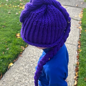 May include: A vibrant purple knitted hat with a braided extension. The hat features a textured knit design and a knotted top. Small gold-colored rings are attached to the braid. The hat is worn with a blue sweatshirt.