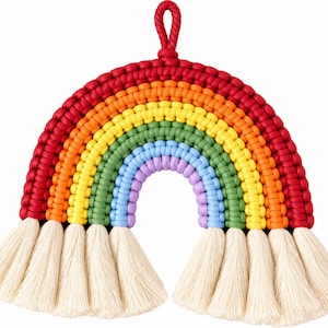 May include: A rainbow-shaped macrame wall hanging constructed from knotted cord in vibrant red, orange, yellow, green, blue, and purple. The bottom of each color arch terminates in a thick, cream-colored cotton tassel fringe with a hanging loop.