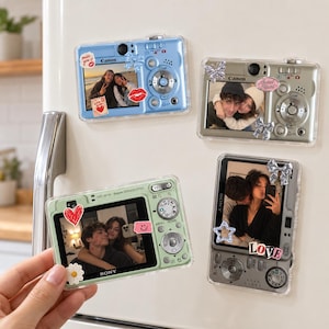 May include: Four rectangular camera-shaped magnets featuring clear plastic covers. Each magnet displays a different digital camera model, including Canon and Sony, with various decorative stickers like hearts, bows, and stars applied over the screens.