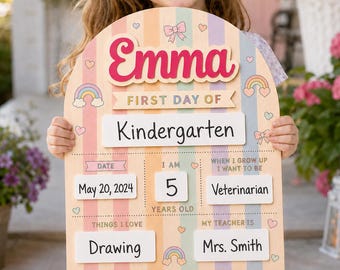 Könnte beinhalten: Ein bogenförmiges Holzschild mit pastellfarbenen vertikalen Streifen und dem Namen Emma in pinker Schreibschrift. Es enthält Felder für First Day of Kindergarten, Date May 20, 2024, Age 5, Veterinarian, Drawing und Teacher Mrs. Smith mit Regenbogen- und Schleifenmotiven.