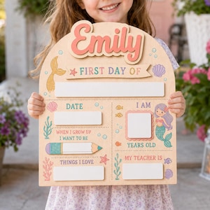 Könnte beinhalten: Ein bogenförmiges Holzschild mit dem Namen Emily in korallenfarbener Schrift, mit Feldern für Datum, Alter, Lehrer und Interessen. Das Schild ist mit Meerjungfrauen-Motiven wie Schwanzflosse, Muscheln, Algen und einem Stift verziert.
