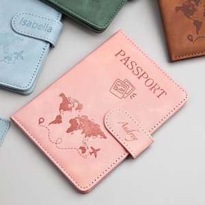 May include: A collection of passport holders in various colors, including pink, blue, green, and brown. The pink passport holder features the word "PASSPORT" and a world map design. The name "Audrey" is on the closure.