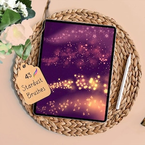 May include: A digital tablet shows a purple and gold stardust design, with a white stylus beside it. A tag reads "43 Stardust Brushes". The tablet is on a woven placemat, with flowers in the background. This is a digital art product.