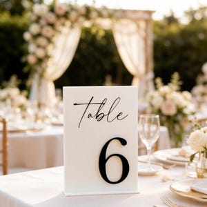 May include: A rectangular white acrylic table number sign featuring the word Table in black cursive script above the large black numeral 6. The sign stands upright on a white tablecloth, displaying a smooth, opaque surface with clean edges.