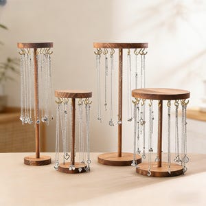 Puede incluir: Cuatro expositores de joyería de madera de diferentes alturas. Cada soporte tiene una parte superior de madera redonda u ovalada con pequeños ganchos para colgar collares. Los soportes sostienen múltiples collares plateados con varios colgantes. Las bases también son de madera.