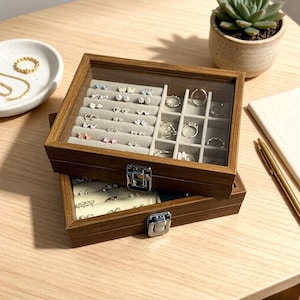 K&ouml;nnte beinhalten: Eine zweist&ouml;ckige Schmuckschatulle aus Holz mit Glasdeckel, die eine Auswahl an Ohrringen und Ringen zeigt. Die Schatulle hat einen hellgrauen Innenraum und einen silbernen Verschluss. Eine kleine Sukkulentenpflanze im Topf ist im Hintergrund zu sehen.