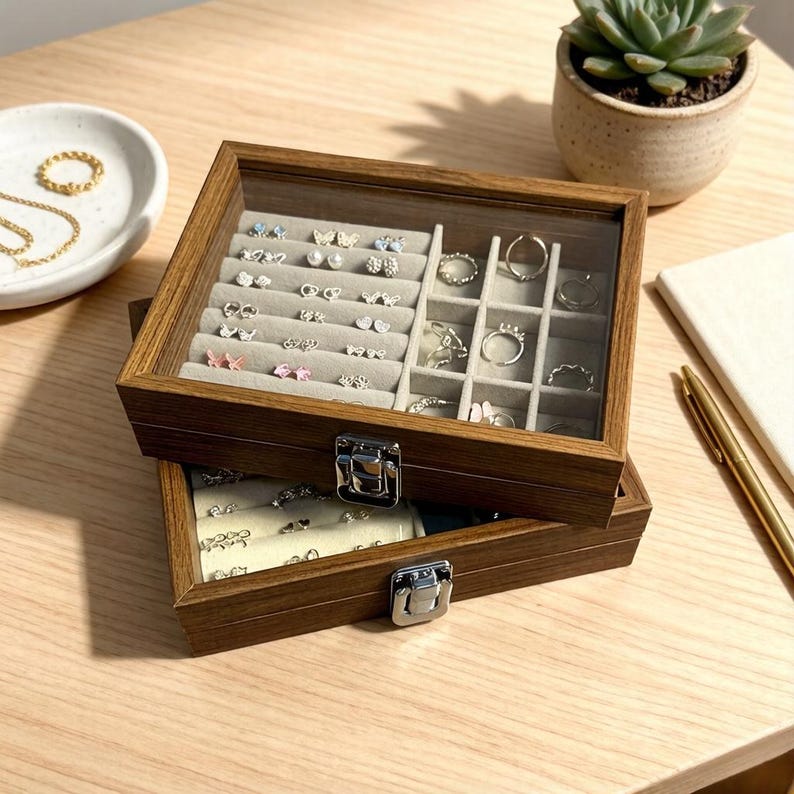 K&ouml;nnte beinhalten: Eine zweist&ouml;ckige Schmuckschatulle aus Holz mit Glasdeckel, die eine Auswahl an Ohrringen und Ringen zeigt. Die Schatulle hat einen hellgrauen Innenraum und einen silbernen Verschluss. Eine kleine Sukkulentenpflanze im Topf ist im Hintergrund zu sehen.