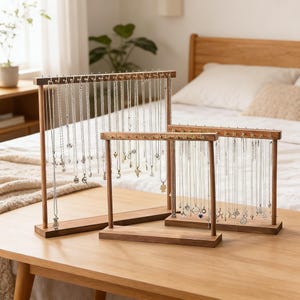 May include: Three wooden jewelry display stands of varying sizes, each with a rectangular base and a top bar with small pegs. The stands hold numerous necklaces with pendants. The wood is a warm brown color, and the necklaces are silver.