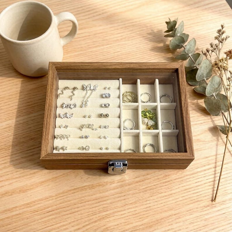K&ouml;nnte beinhalten: Eine Holzschmuckschatulle mit Glasdeckel, gef&uuml;llt mit verschiedenen Ohrringen und Ringen. Die Schatulle ist in F&auml;cher mit beigem Innenraum unterteilt. Links neben der Schatulle steht eine Keramiktasse auf einer Holzoberfl&auml;che.