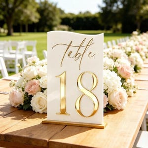 Può includere: Un segnatavolo rettangolare in acrilico bianco con bordo superiore ondulato, caratterizzato dalla scritta dorata "Table" in corsivo sopra il grande numero "18" dorato e riflettente. Il segnatavolo è sostenuto da una sottile base dorata.