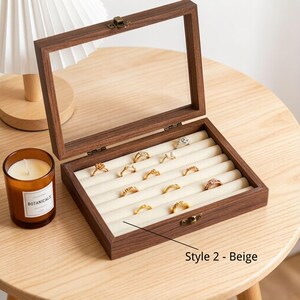 K&ouml;nnte beinhalten: Eine braune Schmuckschatulle aus Holz mit einem durchsichtigen Glasdeckel, ge&ouml;ffnet, um Ringe auf beigen Samtrollen zu pr&auml;sentieren. Die Schatulle hat einen Messingverschluss. Der Text "Style 2 - Beige" ist sichtbar.