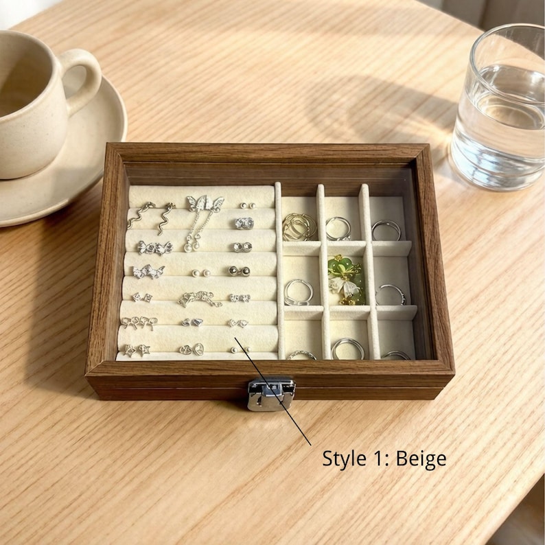 K&ouml;nnte beinhalten: Eine Holzschmuckschatulle mit Glasdeckel, gef&uuml;llt mit Ohrringen und Ringen. Der Innenraum ist beige mit mehreren F&auml;chern. Die Schatulle wird mit einem silbernen Verschluss verschlossen. Der Text "Style 1: Beige" ist sichtbar.