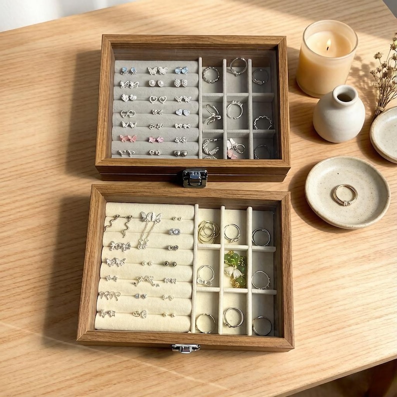 K&ouml;nnte beinhalten: Zwei offene Schmuckk&auml;stchen aus Holz mit Glasdeckeln, die eine Auswahl an Ohrringen und Ringen zeigen. Die obere Box hat einen grauen Innenraum, die untere einen beigen. Eine brennende Kerze, eine kleine Vase und ein Ring auf einem Teller sind ebenfalls zu sehen.