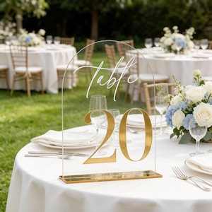 May include: A clear acrylic arched table number sign featuring the white script text Table and the large gold numeral 20. The sign is supported by a rectangular gold base and is positioned on a white tablecloth with place settings.