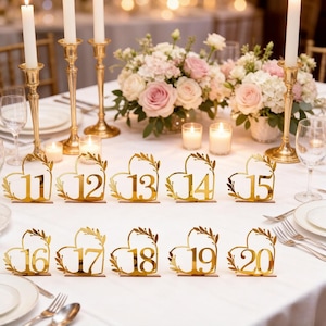 May include: Gold heart-shaped table numbers 11-20 with leaf accents. The numbers are displayed on a white tablecloth, with candles, floral arrangements, and place settings in the background. Ideal for weddings or special events.