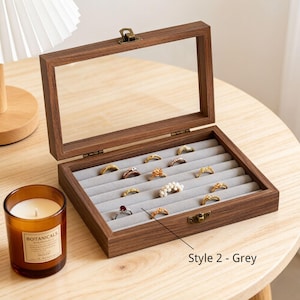 K&ouml;nnte beinhalten: Eine Holzschmuckschatulle mit Glasdeckel, ge&ouml;ffnet, um verschiedene Ringe auf einem grauen Samtinnenraum zu pr&auml;sentieren. Die Schatulle ist braun mit einem Messingverschluss. Eine Kerze in einem Glas steht daneben.