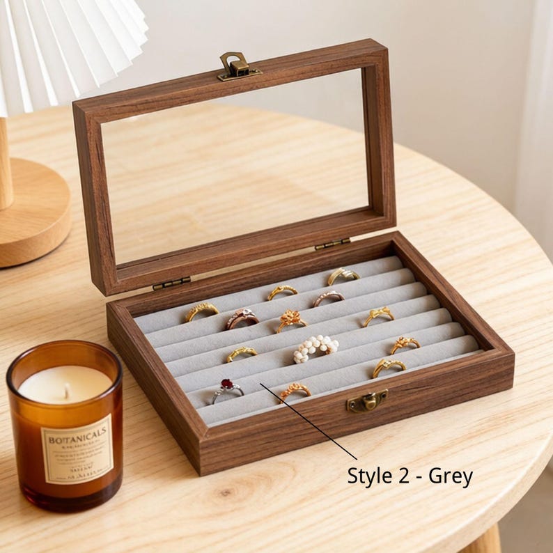 K&ouml;nnte beinhalten: Eine Holzschmuckschatulle mit Glasdeckel, ge&ouml;ffnet, um verschiedene Ringe auf einem grauen Samtinnenraum zu pr&auml;sentieren. Die Schatulle ist braun mit einem Messingverschluss. Eine Kerze in einem Glas steht daneben.