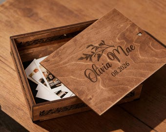 May include: A wooden keepsake box with a hinged lid, partially open to reveal photographs. The lid is engraved with the name "Olivia Mae" and the date "08.2026", along with a floral design. The box is made of light brown wood.