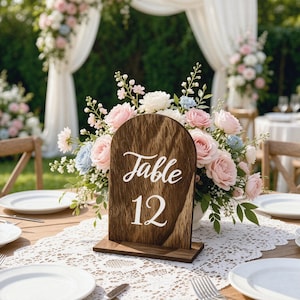 May include: An arched wooden table number sign with a dark wood grain finish, featuring the white text Table 12 in a script and serif font. The sign is positioned in front of a floral arrangement with pink and light blue roses and greenery.