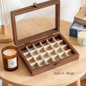 K&ouml;nnte beinhalten: Eine Holzschmuckschatulle mit einem Deckel aus klarem Glas und einem Messingverschluss. Der Innenraum ist in kleine F&auml;cher mit beigem Stofffutter unterteilt, die mehrere goldene Ringe enthalten. Die Box ist mit "Style 4 - Beige" beschriftet.