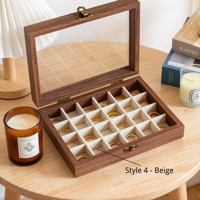 K&ouml;nnte beinhalten: Eine Holzschmuckschatulle mit einem Deckel aus klarem Glas und einem Messingverschluss. Der Innenraum ist in kleine F&auml;cher mit beigem Stofffutter unterteilt, die mehrere goldene Ringe enthalten. Die Box ist mit "Style 4 - Beige" beschriftet.
